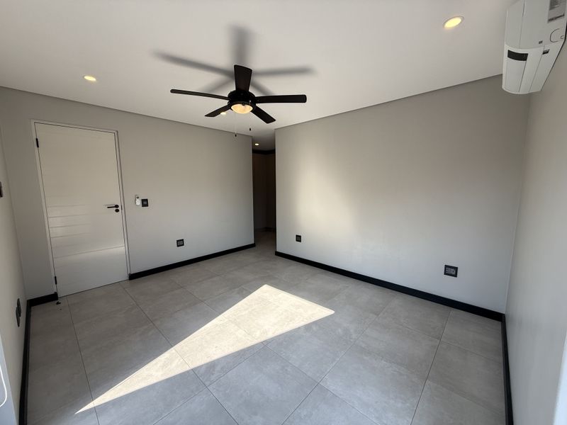 A bright, modern, and empty bedroom featuring neutral tiled flooring, recessed lighting, a ceiling fan, and a wall-mounted air conditioning unit.