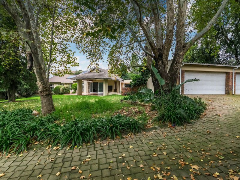 Front view of the property featuring a paved driveway, double garage, and a covered patio entrance surrounded by mature trees and garden space.