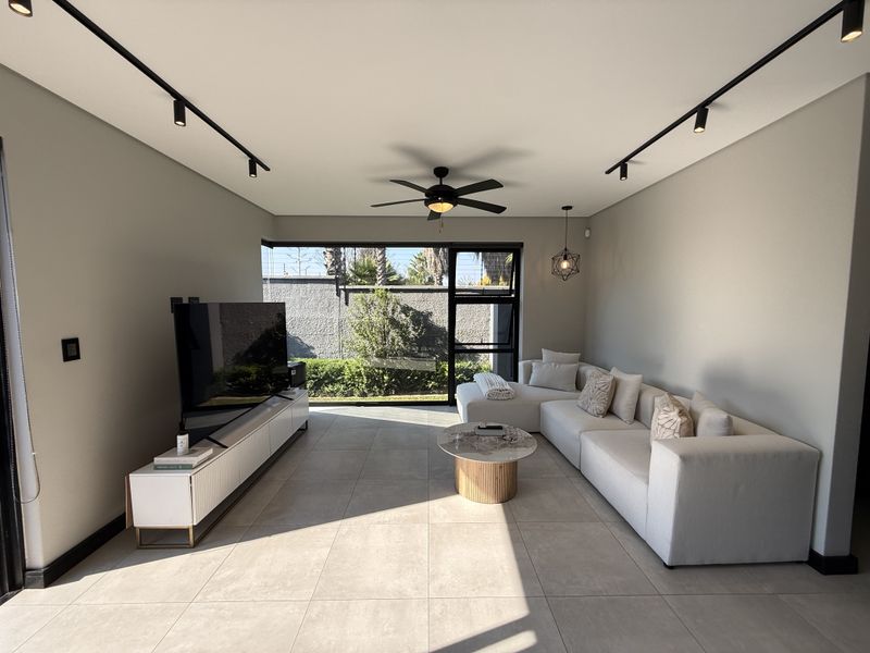 Modern living room featuring large format floor tiles, track lighting, a ceiling fan, and a large corner window overlooking the garden.