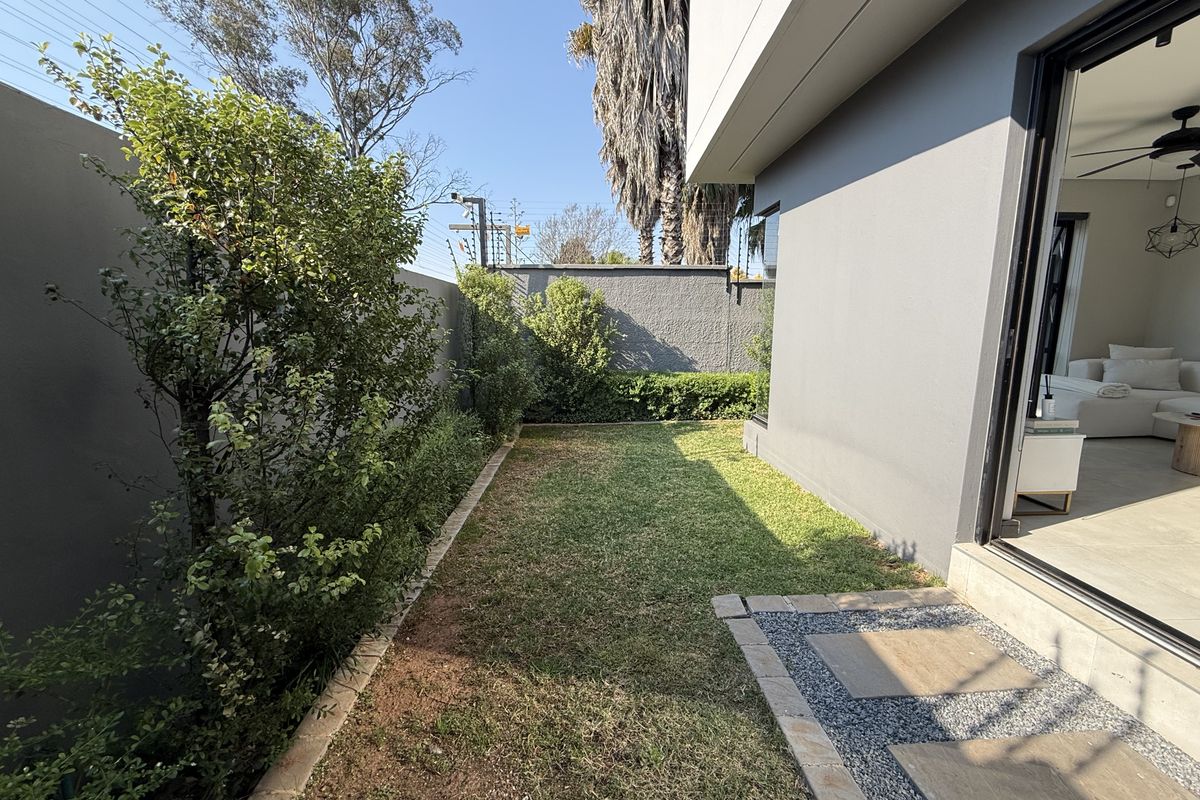 A private rear garden featuring a manicured lawn, boundary walls, and a paved walkway leading to the main living area through sliding glass doors.