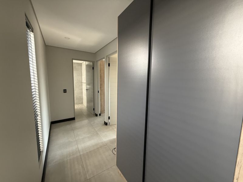 A modern, well-lit hallway featuring neutral tiled flooring, built-in cupboards, and clean finishes leading to adjacent rooms.