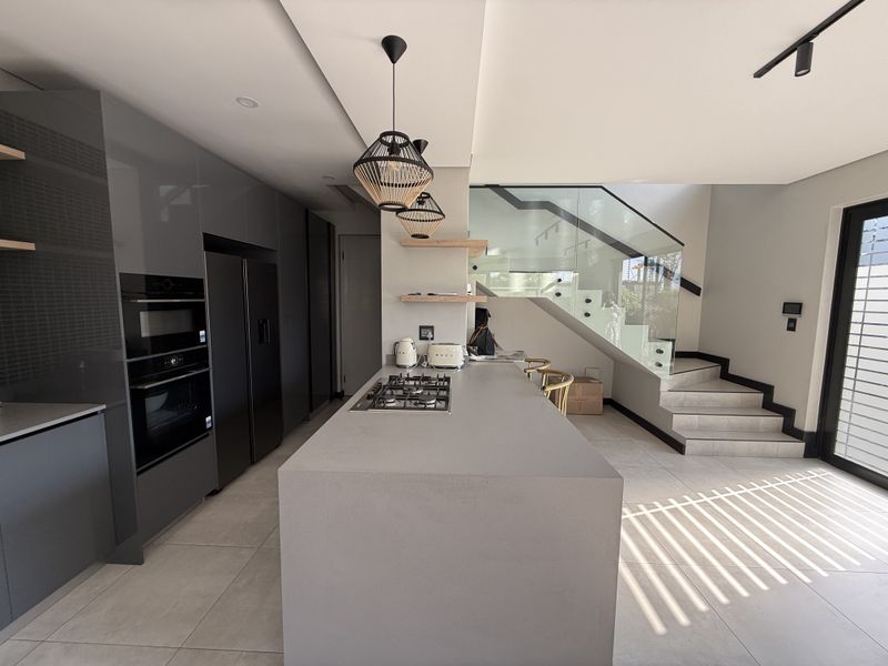 Modern open-plan kitchen featuring a central island with gas hob, sleek dark cabinetry, built-in appliances, and a contemporary staircase with glass balustrades.