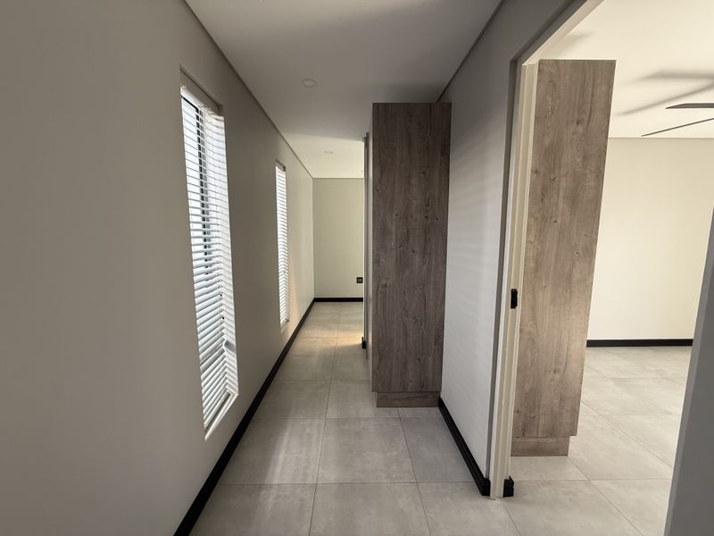 A modern, well-lit hallway featuring neutral tiled flooring, built-in wood-finish cabinetry, and clean lines, leading into an adjacent living space.