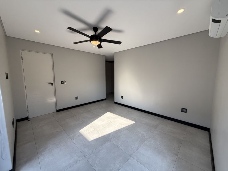 A bright, modern, and empty bedroom featuring neutral tiled flooring, a ceiling fan, and a wall-mounted air conditioning unit.