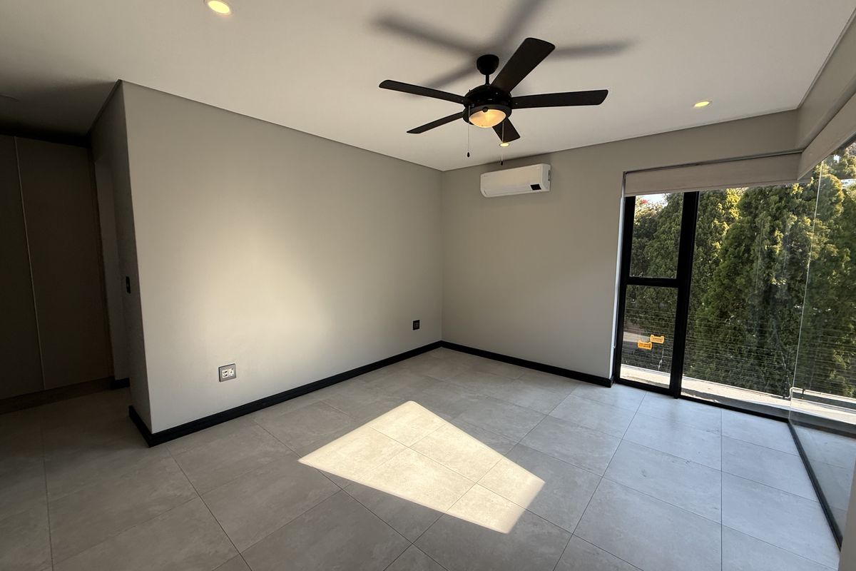 A bright, modern bedroom featuring neutral tiled flooring, a ceiling fan, split-system air conditioning, and large windows with views of surrounding greenery.