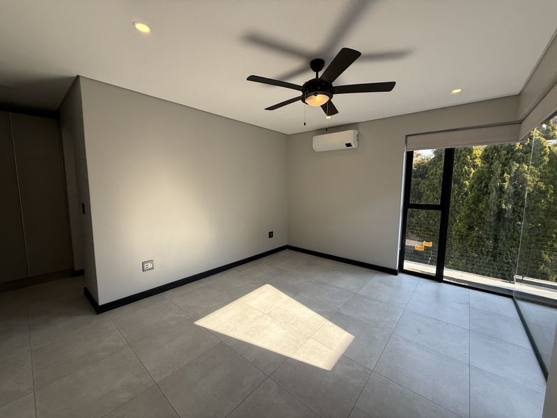 A bright, modern bedroom featuring neutral tiled flooring, a ceiling fan, split-system air conditioning, and large windows with views of surrounding greenery.