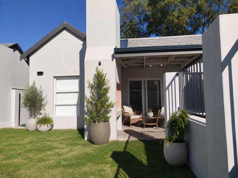 A well-maintained rear exterior featuring a paved patio, built-in braai, and sliding glass doors leading into the home, set against a landscaped garden.