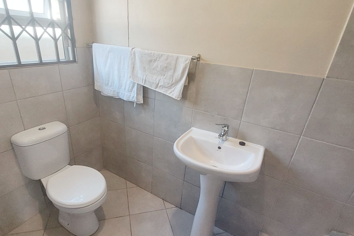 A functional bathroom featuring a pedestal basin, toilet, and neutral wall and floor tiling with a security-barred window.