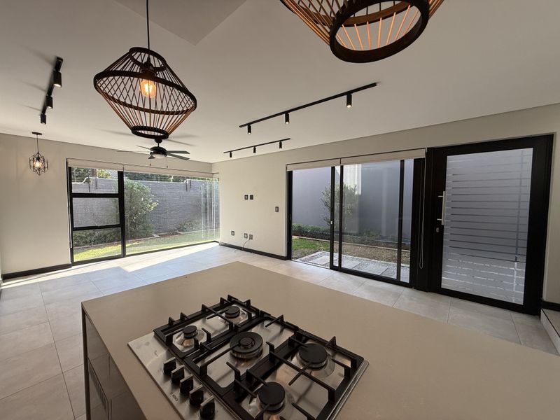 Modern open-plan living and kitchen area featuring a central island with gas hob, sleek track lighting, and large sliding doors opening to the garden.