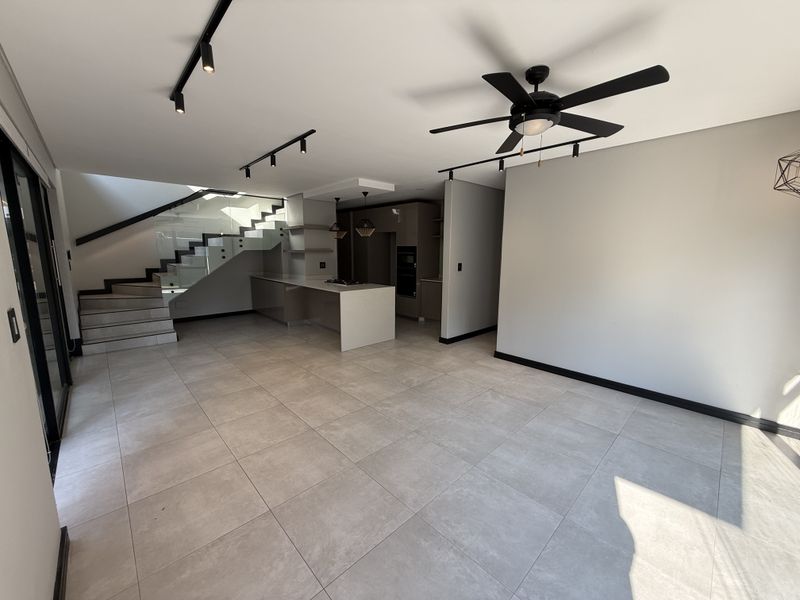 Modern open-plan living area featuring a sleek kitchen with island, contemporary track lighting, ceiling fan, and a staircase with a glass balustrade.