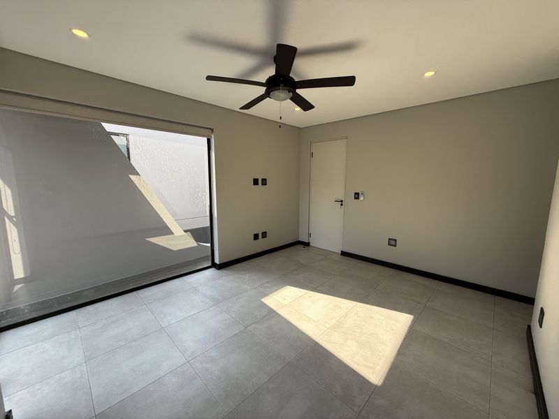 A bright, modern, and empty bedroom featuring neutral tiled flooring, a ceiling fan, recessed lighting, and a large sliding glass door leading outdoors.
