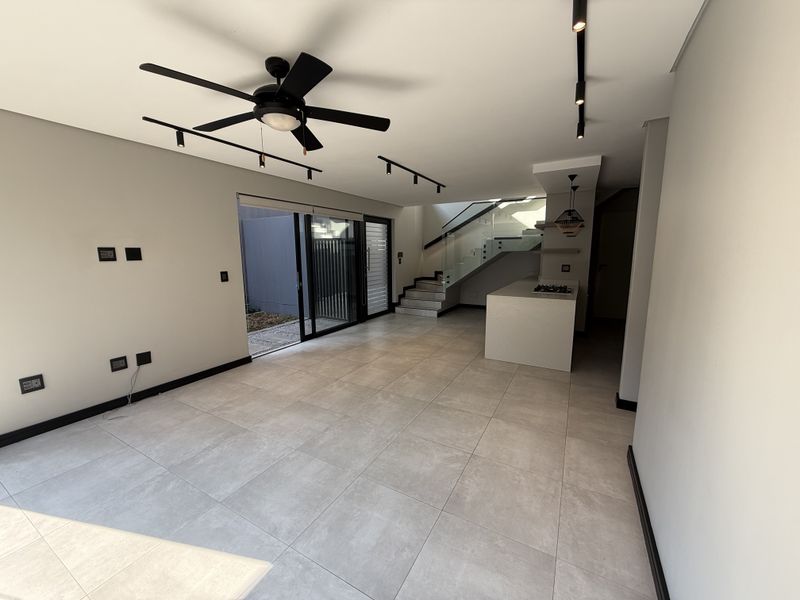 Modern open-plan living area featuring large format floor tiles, track lighting, a sleek kitchen island with a gas hob, and a glass-railed staircase.