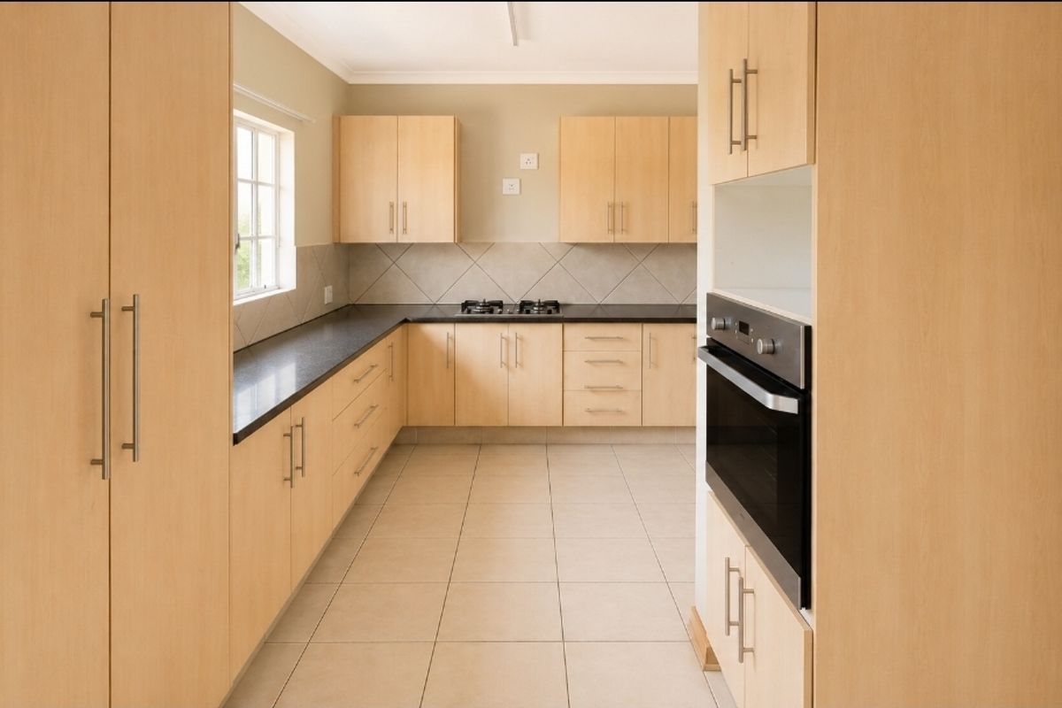 A clean, functional kitchen featuring light wood-finish cabinetry, dark countertops, a built-in oven, and a gas hob with neutral tiled flooring.