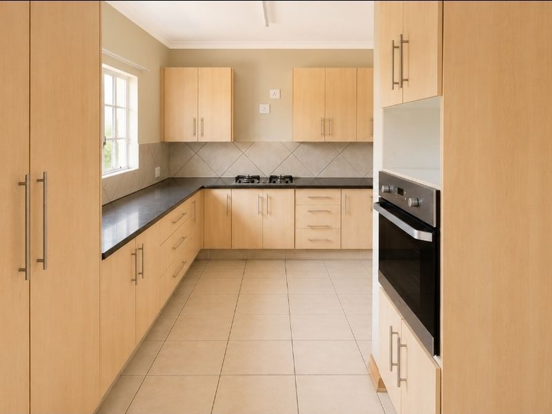 A clean, functional kitchen featuring light wood-finish cabinetry, dark countertops, a built-in oven, and a gas hob with neutral tiled flooring.