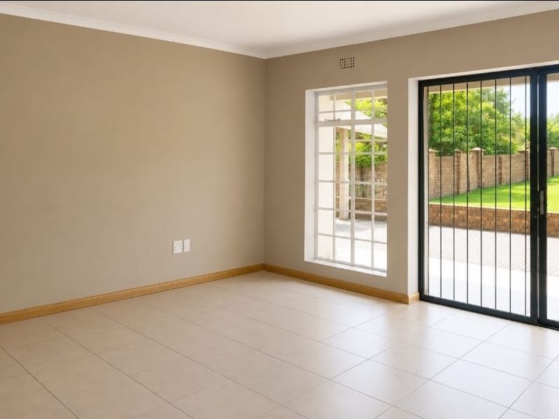 A bright, empty living room featuring neutral tiled flooring, large windows, and a sliding glass door with security bars leading to the garden.