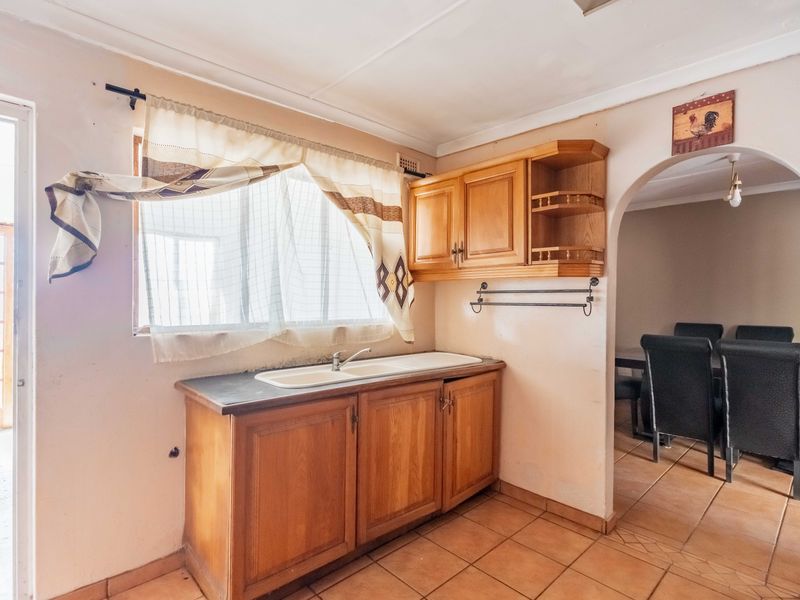 A dated kitchen area featuring wooden cabinetry, a double basin sink, and tiled flooring, with an arched opening leading to the adjacent dining space.