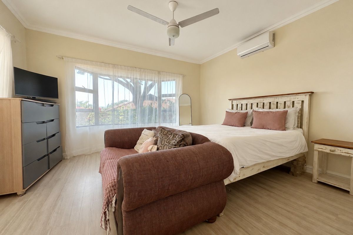 A well-lit bedroom featuring laminate flooring, a ceiling fan, and a split-system air conditioner, with a large window providing natural light.