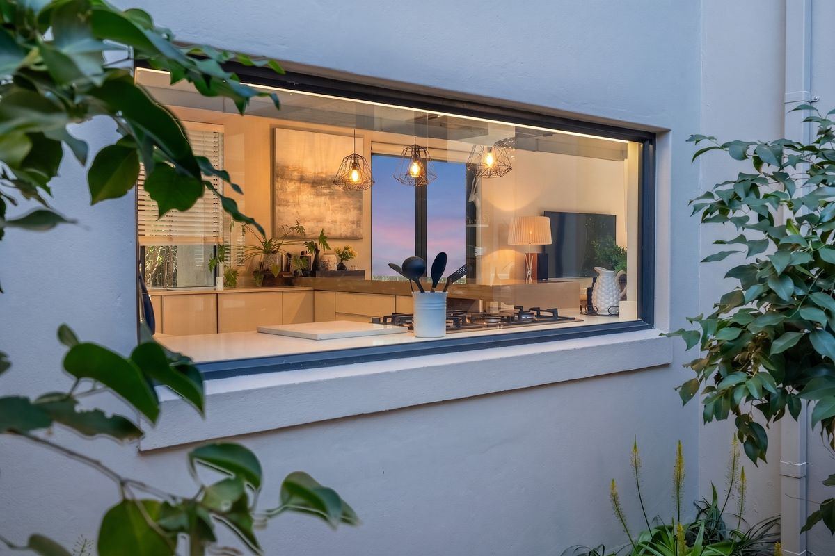 A modern kitchen interior viewed through a large exterior window, featuring sleek cabinetry, stone countertops, and a built-in gas hob.