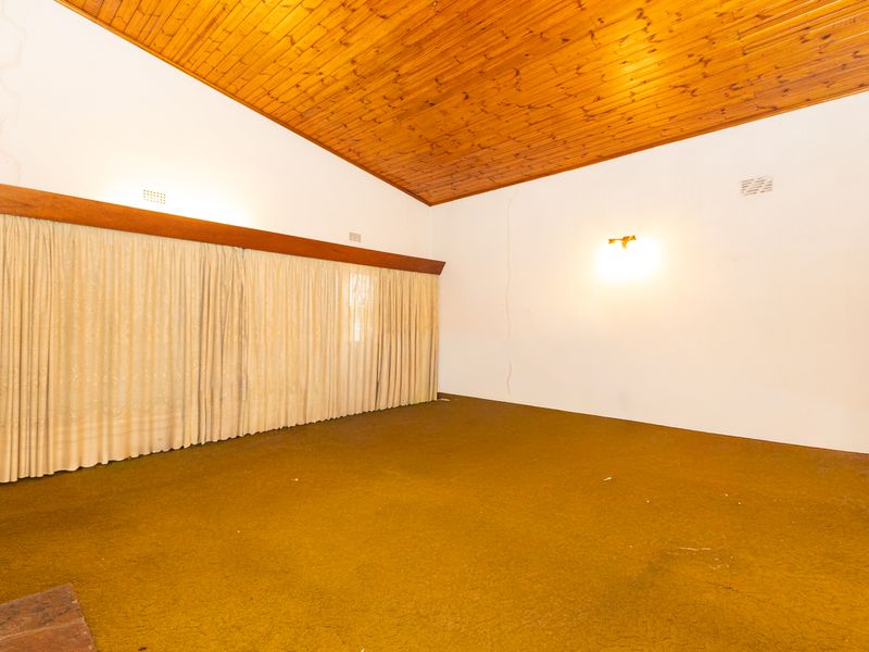 An empty living room featuring a high raked timber-clad ceiling, wall-mounted lighting, and carpet flooring, requiring cosmetic updates to the walls and finishes.