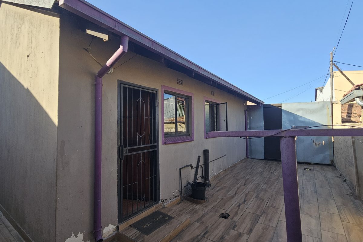 Rear exterior view of the property featuring a tiled patio area, security gate, and basic wall finishes requiring maintenance.