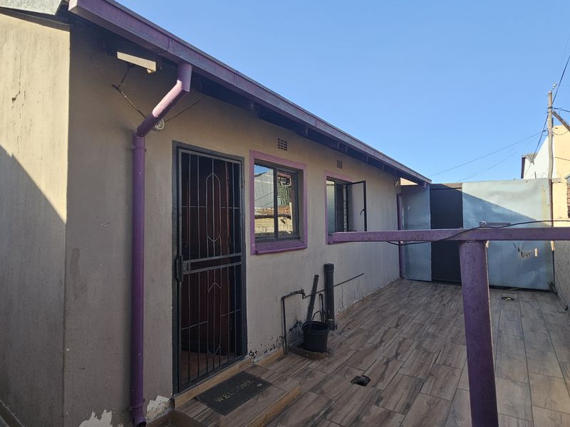 Rear exterior view of the property featuring a tiled patio area, security gate, and basic wall finishes requiring maintenance.