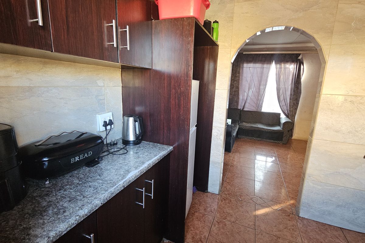 A compact kitchen featuring dark wood cabinetry, granite-look countertops, and tiled finishes, leading through an arched doorway into the adjacent living area.