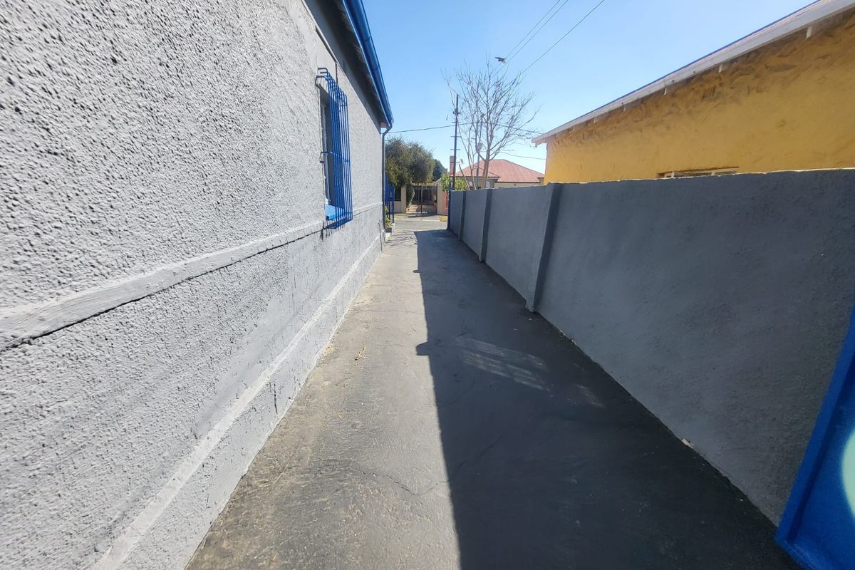 A narrow paved side passage between two residential buildings, featuring a grey boundary wall and security bars on the window.