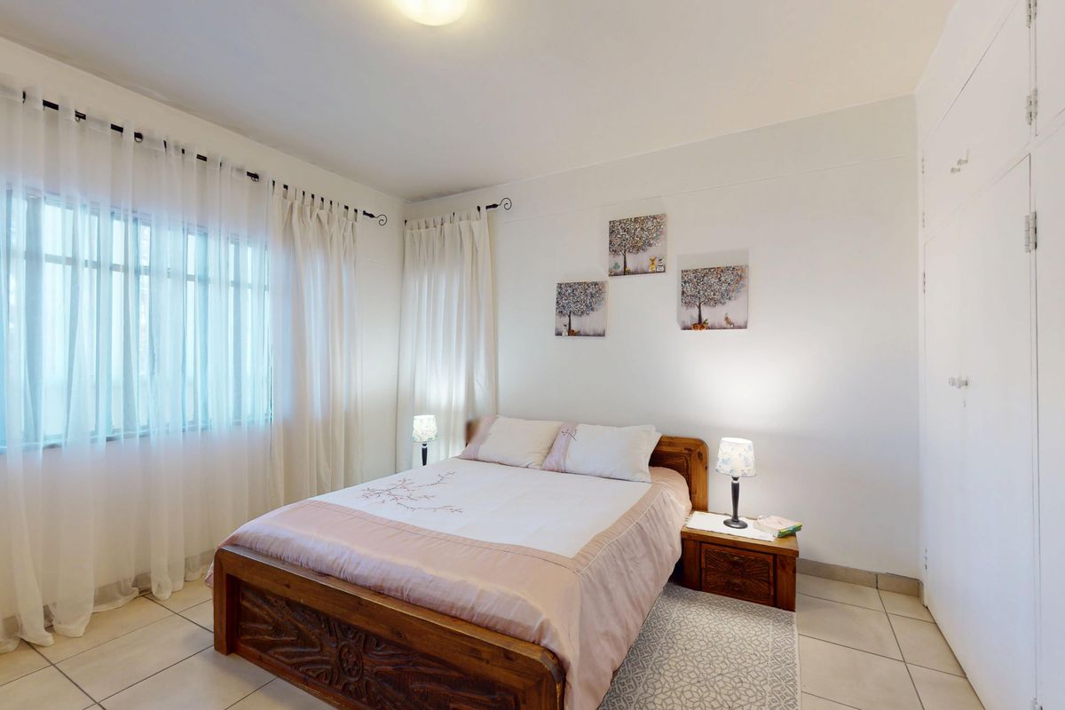 A bright bedroom featuring tiled flooring, built-in cupboards, and large windows with curtains, offering a clean and functional living space.