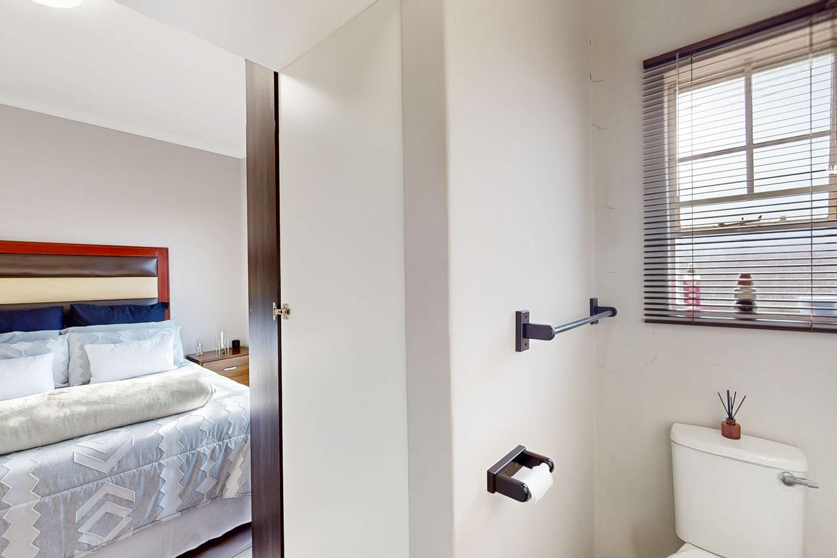 A functional bathroom space featuring a toilet, wall-mounted fixtures, and a window with blinds, adjacent to a bedroom area.