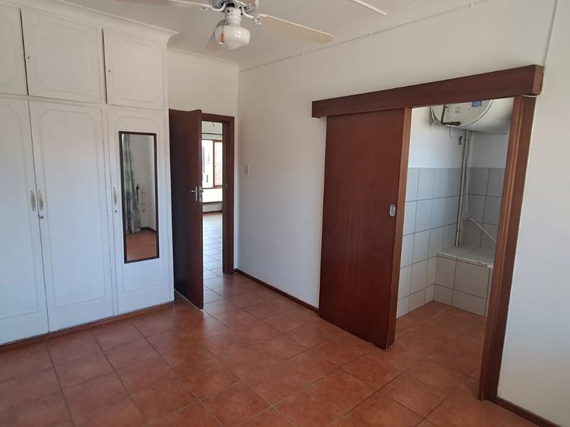 An empty bedroom featuring built-in cupboards, tiled flooring, a ceiling fan, and an adjacent tiled utility area with a geyser.