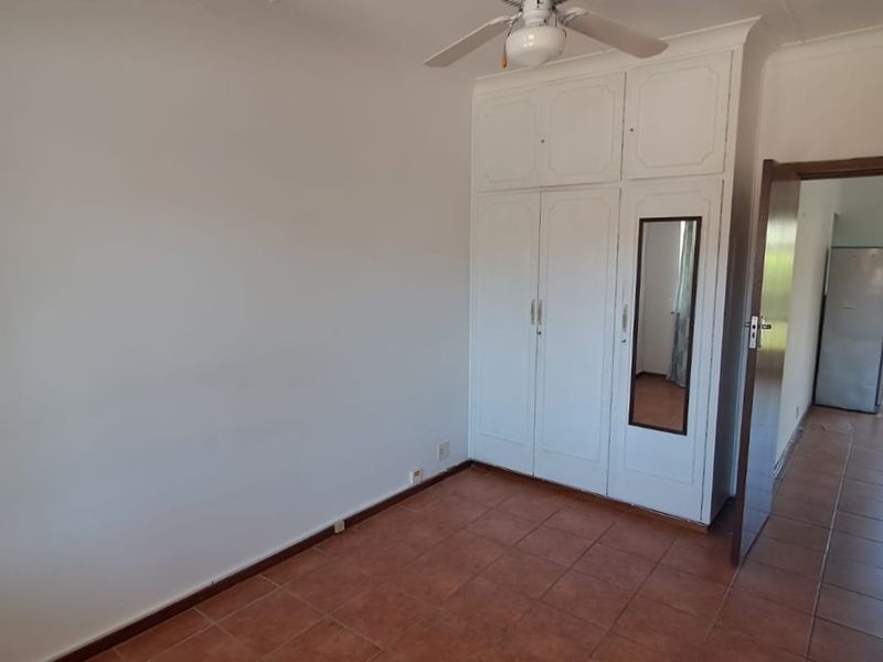 A bright, empty bedroom featuring built-in cupboards with an integrated mirror, a ceiling fan, and durable tiled flooring.
