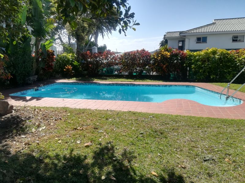 A well-maintained outdoor swimming pool area featuring brick coping, a metal ladder, and surrounding landscaped garden beds.