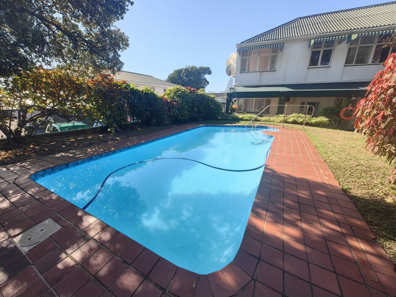 A well-maintained outdoor swimming pool area featuring brick paving, a safety handrail, and established garden landscaping within the retirement village grounds.