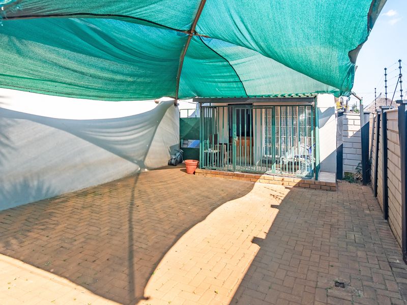 A paved outdoor patio area featuring a large shade cloth canopy, enclosed by perimeter walls and secured with a metal gate.