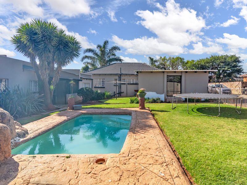 A sunny backyard featuring a swimming pool with a stamped concrete surround, a well-maintained lawn, and established garden trees.