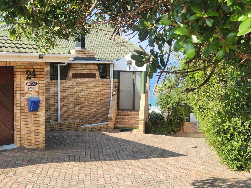 Front exterior view of a brick house featuring a paved driveway, garage, and tiled roof, with mature garden landscaping and a glimpse of the coast.