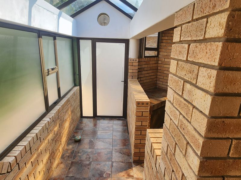 A covered outdoor patio area featuring exposed brickwork, tiled flooring, and a glass roof, providing a versatile space for outdoor living.