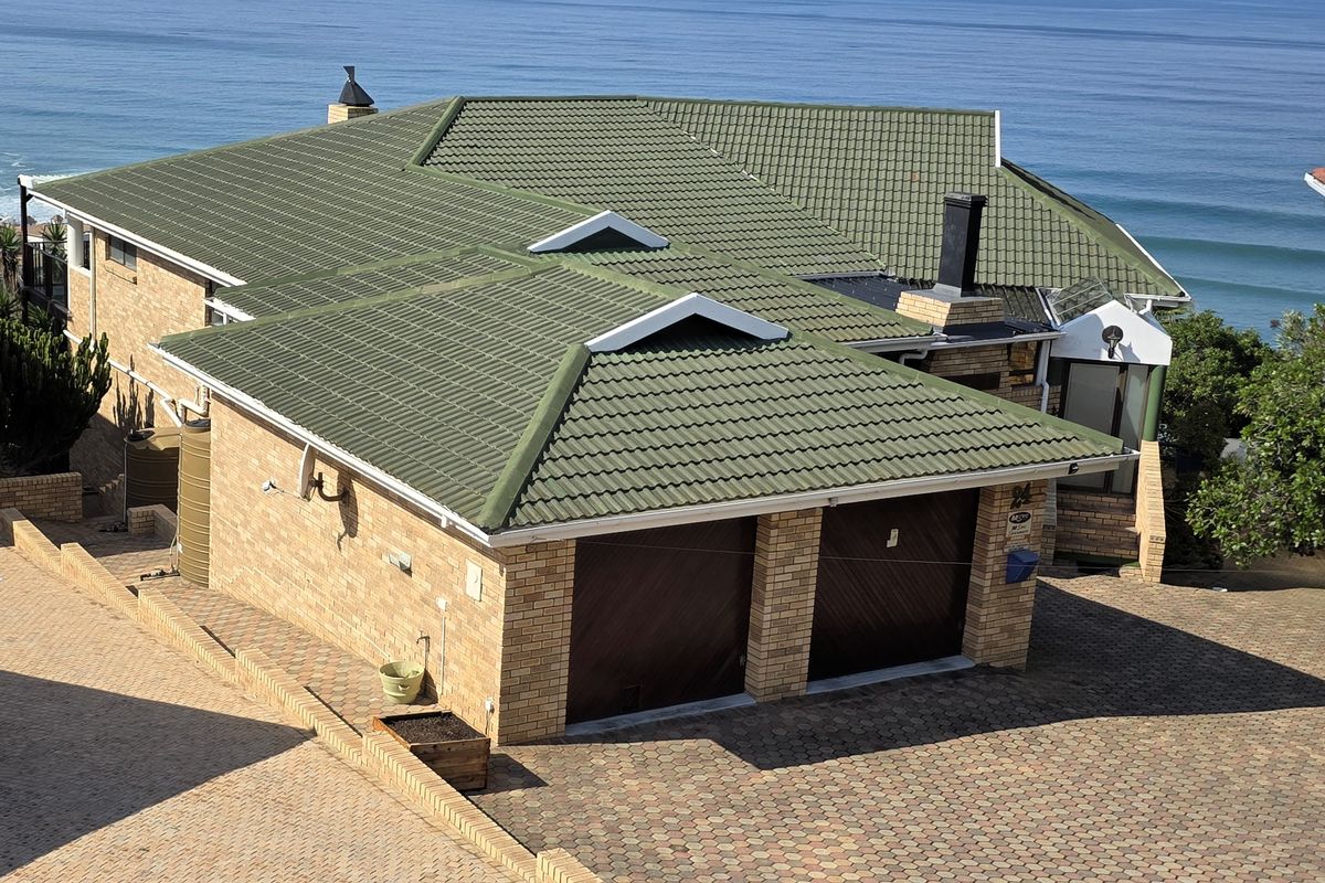 Aerial view of a brick house featuring a green tiled roof, double garage, and paved driveway, situated with a coastal ocean backdrop.