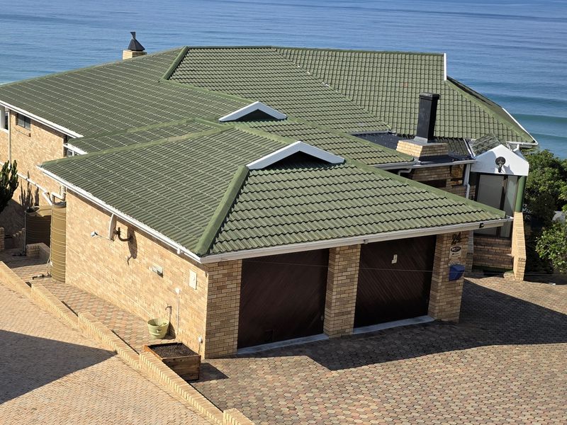 Aerial view of a brick house featuring a green tiled roof, double garage, and paved driveway, situated with a coastal ocean backdrop.