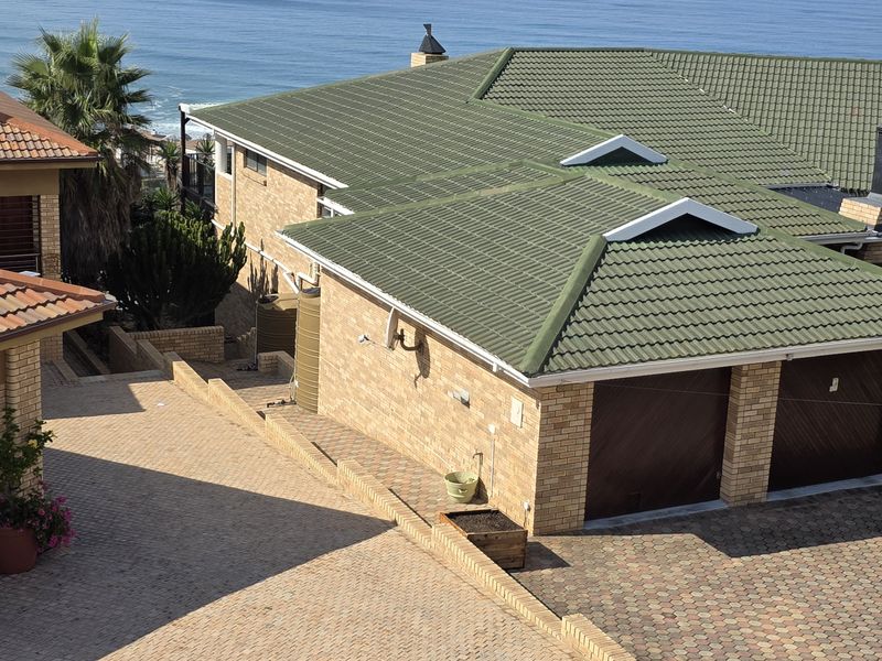 Aerial view of a coastal property featuring brick facades, tiled roofs, double garages, and paved driveways with ocean views in the background.