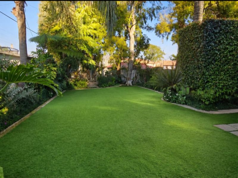 A well-maintained private garden featuring a lush artificial lawn, stone pathway, and established mature trees and greenery.