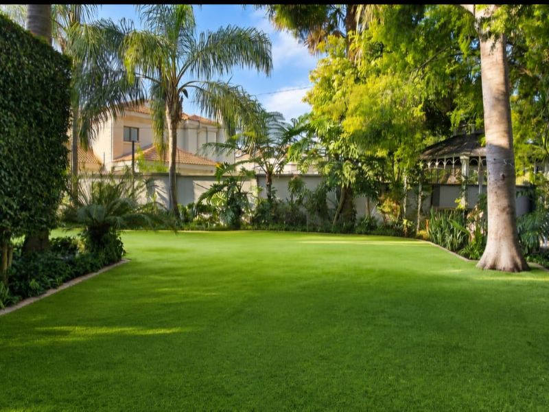 A well-maintained, lush green garden featuring mature trees, manicured hedging, and a secure perimeter wall.