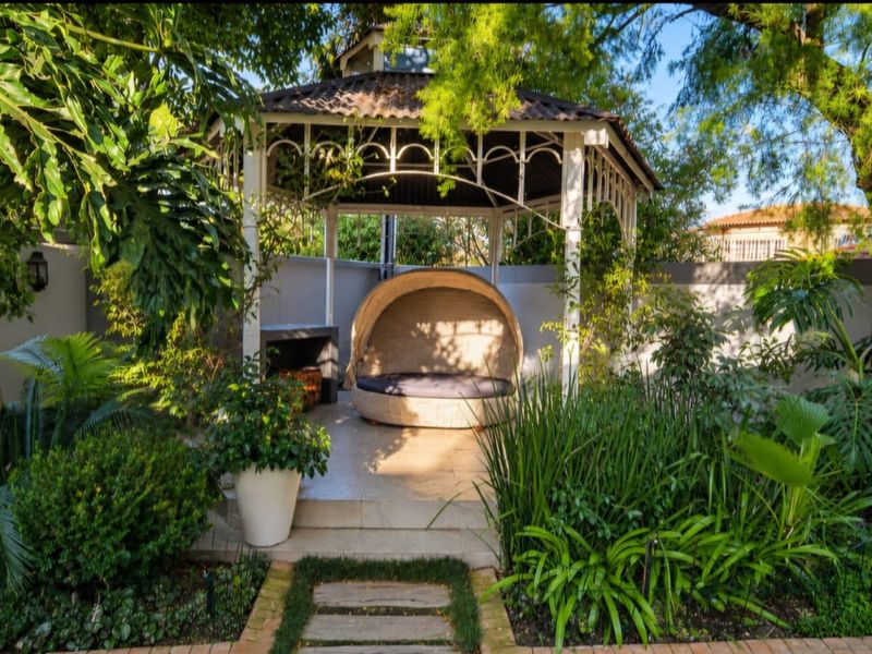 A tranquil garden setting featuring a covered gazebo with tiled flooring and a built-in braai, surrounded by lush landscaping and mature trees.