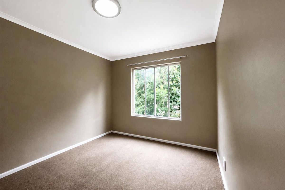A clean, empty bedroom featuring neutral-toned walls, carpeted flooring, and a window providing natural light and garden views.