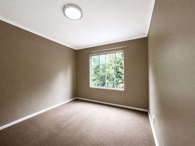 A clean, empty bedroom featuring neutral-toned walls, carpeted flooring, and a window providing natural light and garden views.