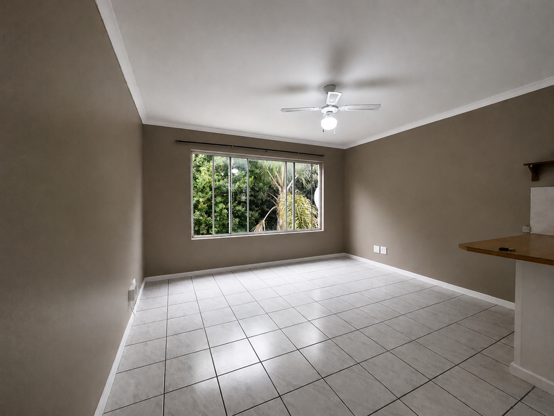 An open-plan living area featuring neutral wall tones, tiled flooring, a ceiling fan, and a breakfast bar overlooking a garden view.
