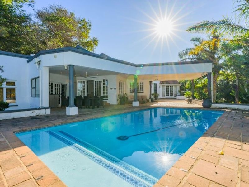 A spacious outdoor pool area featuring a large swimming pool, paved decking, and a covered patio space ideal for outdoor entertaining.