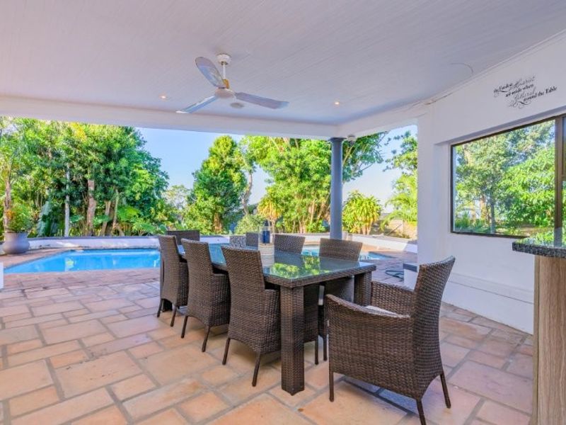 Covered outdoor patio featuring stone tile flooring, a ceiling fan, and a built-in bar counter, overlooking a private swimming pool and garden area.