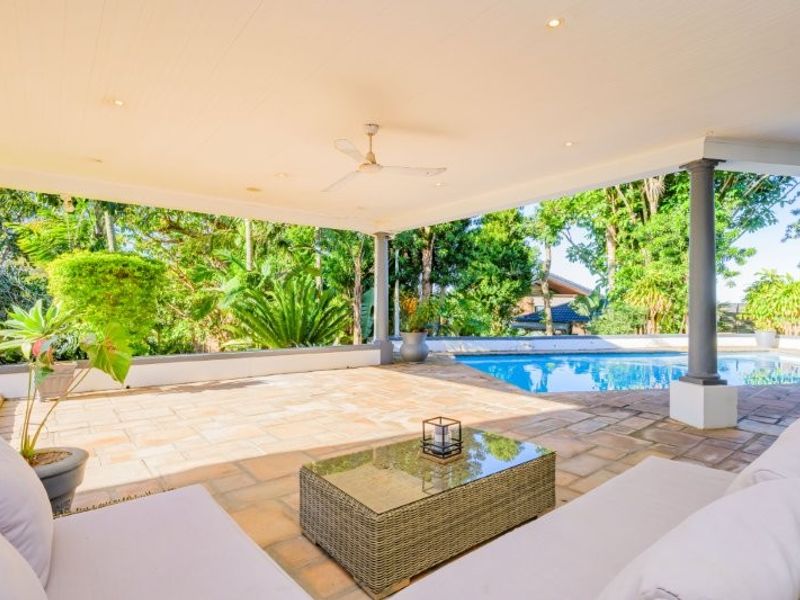 Spacious covered patio featuring stone flooring, recessed lighting, and a ceiling fan, overlooking a private swimming pool and lush garden area.