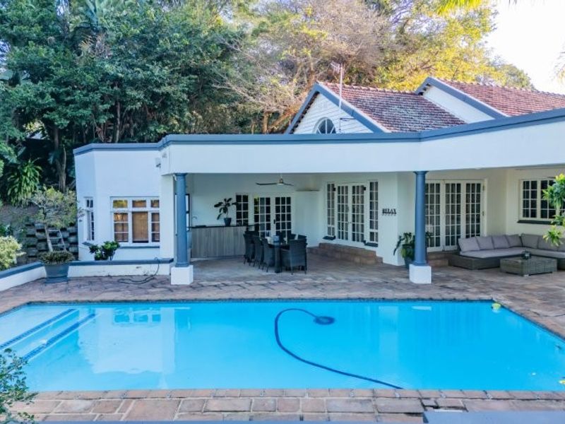 Spacious covered patio overlooking a well-maintained swimming pool, featuring brick paving, built-in bar area, and multiple sets of french doors leading indoors.